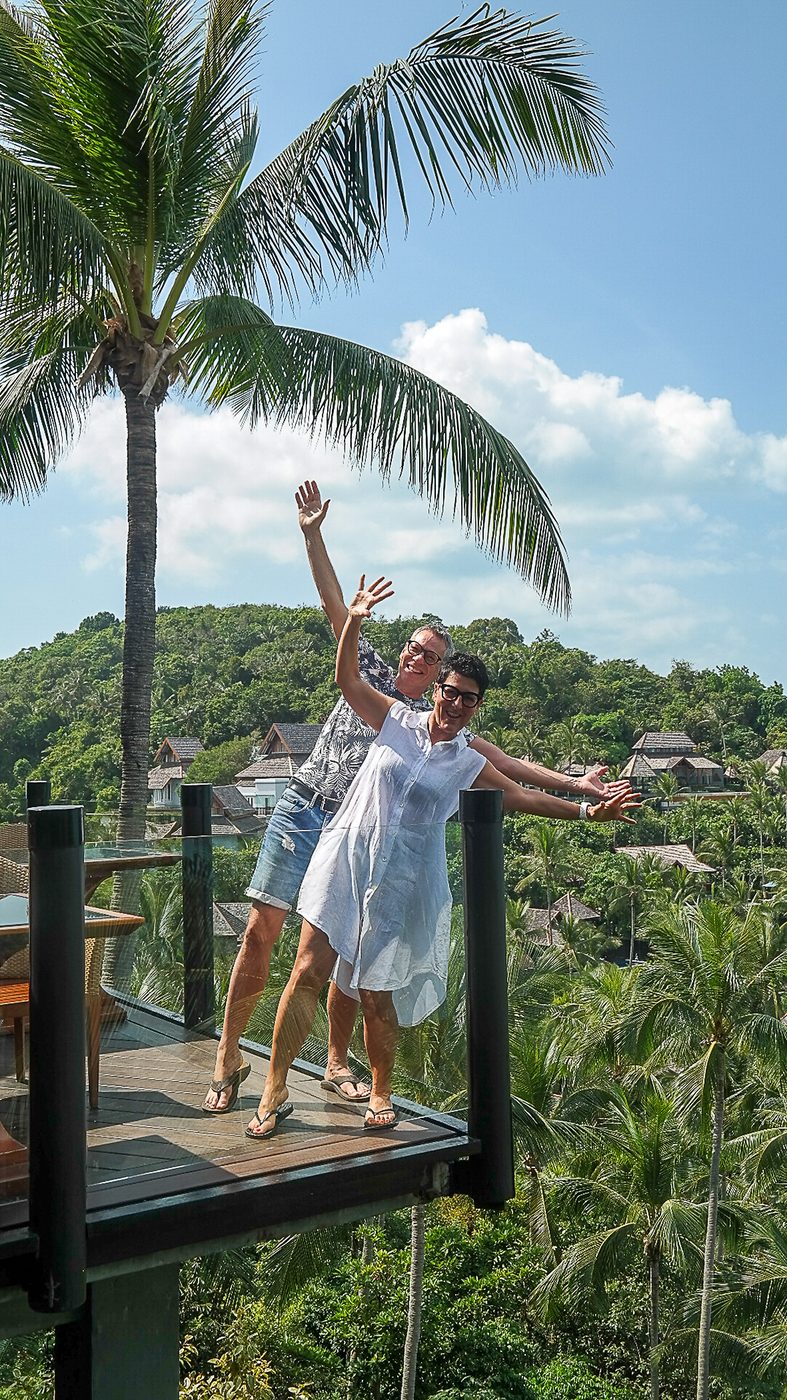 Two guests waving from a treetop deck above a palm-covered hillside