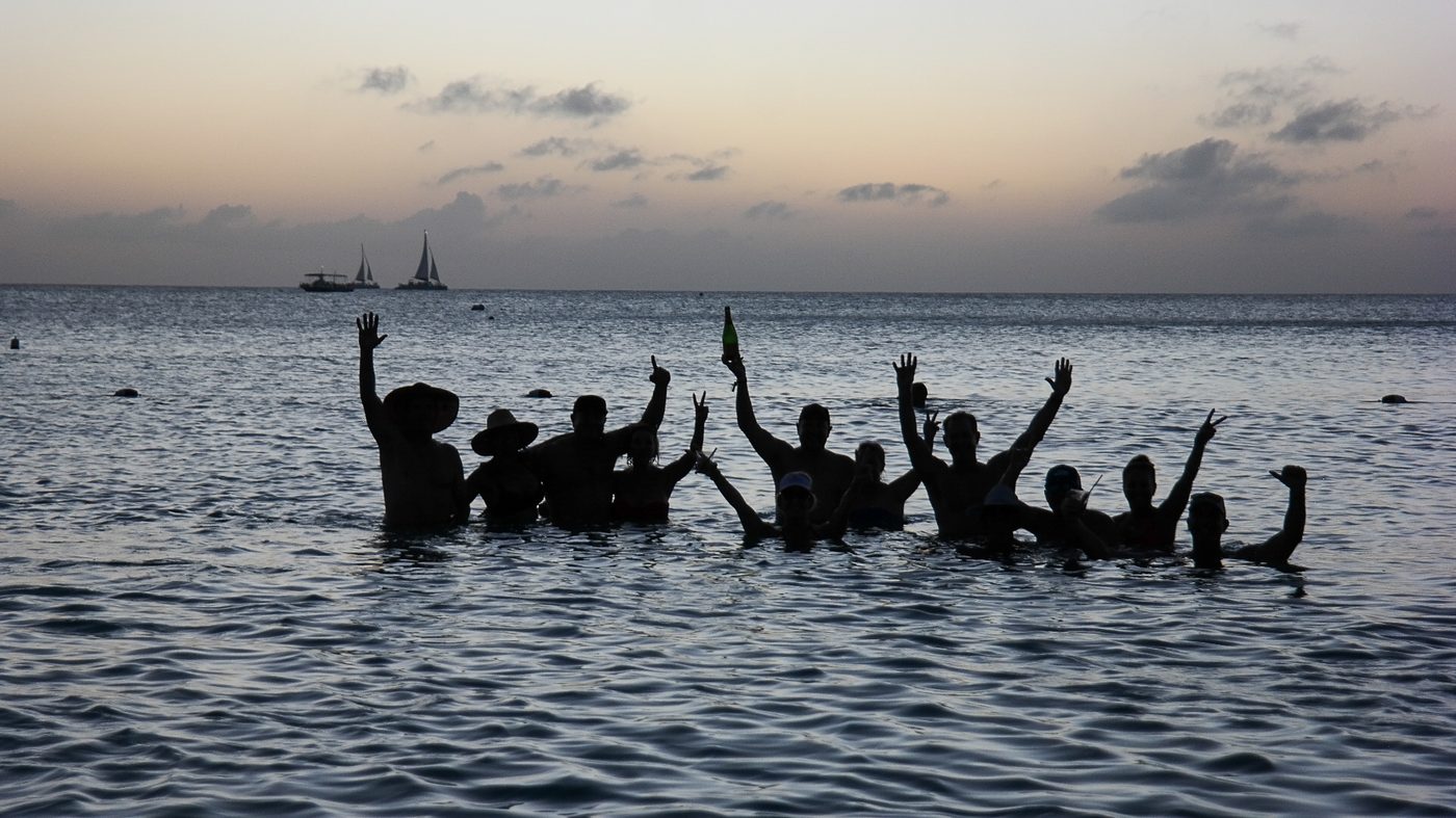 A group standing in shallow ocean water at sunset, hands raised